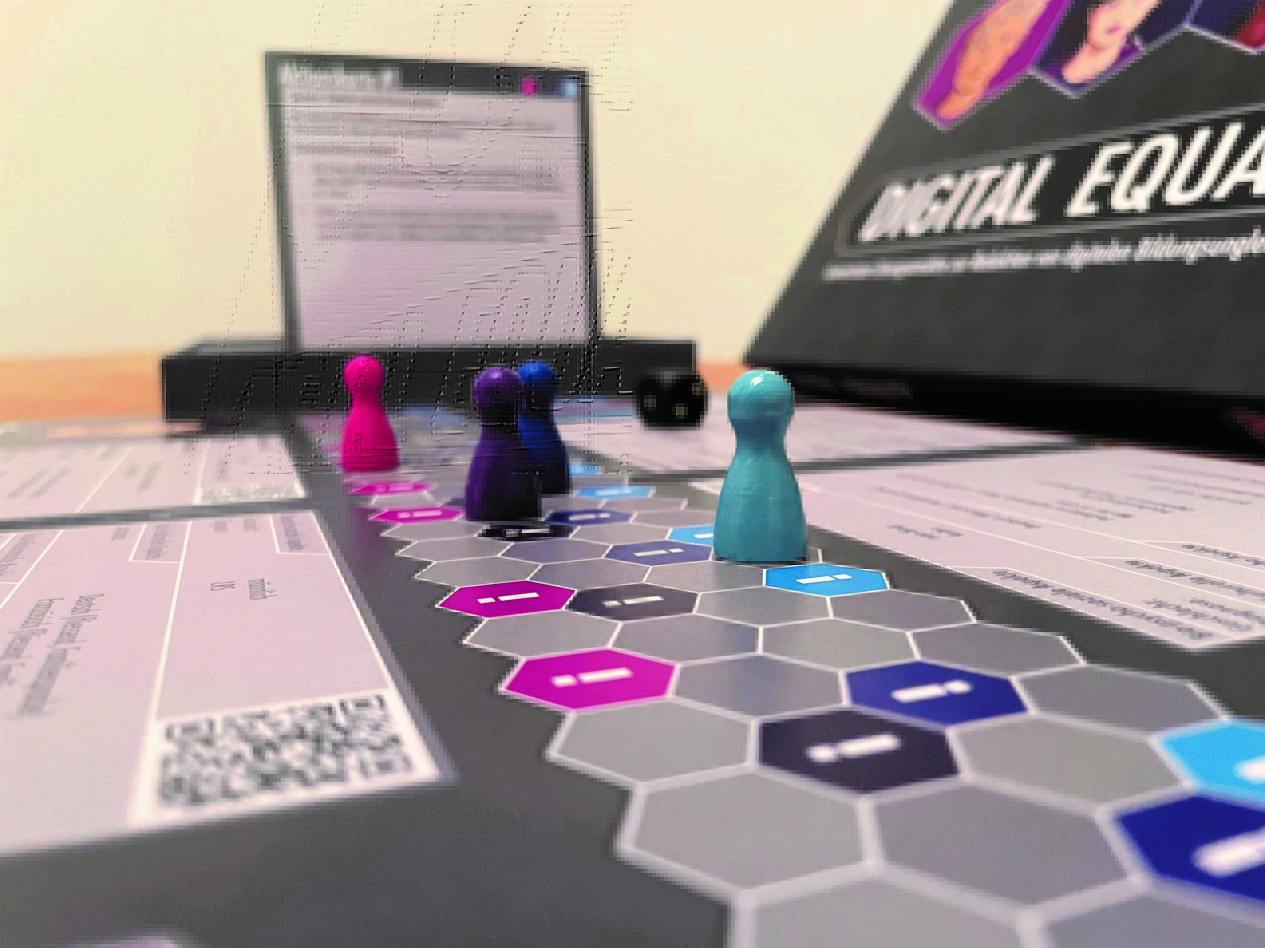Ein Spielfeld aus Sechsecken, die aneinandergelegt sind und jeweils eine Linie oder eine Art Leiter bilden. Vier Spielfiguren sind jeweils auf die vier Leitern verteilt und stehen an unterschiedlichen Positionen. Im Hintergrund ist eine grosse Spielkarte mit Beschreibung und auf der rechten Seite ein Spielbox auf der steht "Digital Equality".